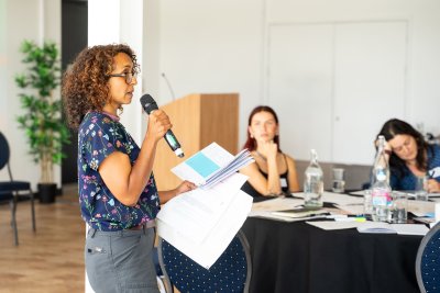 Dr Nicola Scott facilitating the decolonial decision making workshop . Nicola is a black worman with curly, brown shoulder length hair, wearing a navy, floral printed top. She is holding some papers and speaking into a microphone. 2 women are sitting at a table in the background listening and taking notes. 