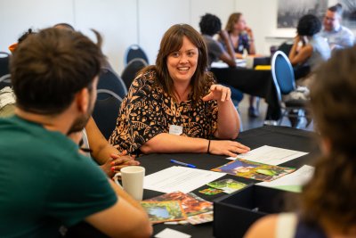 Participant at the 'Embodying the power of food identities' workshop at the Gathering Table 2025. A white woman with shoulder length brown hair wearing a navy top with an orange pattern is sitting at a table with 3 other people, smiling and talking. Pictures and papers are scattered on the table top.