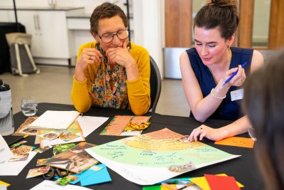 Particpants at the Embodying the power of food identities workshop. 2 women are smiling and looking at the pictures and coloured paper on the table